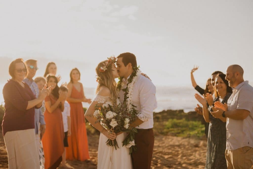 Couple kisses on the beach with friends and family cheering them on in the background at sunset