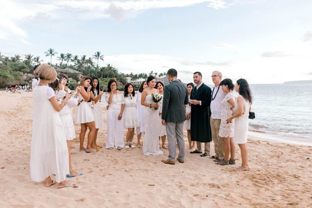 Couple exchanges vows on a tropical beach with all their guests dressed in white, standing in a U shape around them.