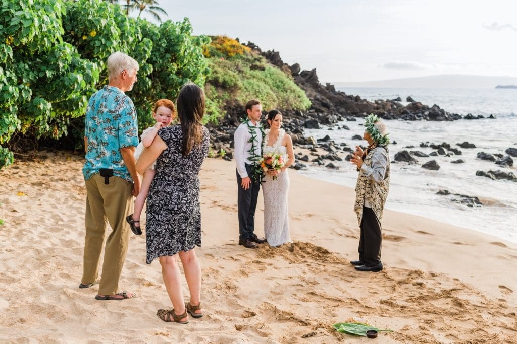 Couple stands side by side on the beach listening to their officiant while their 3 guests watch from the side. One woman holds a child.
