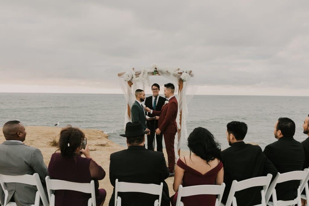 Gay couple stands in front of an arch with their officiant in view of the ocean with their friends and family watching the elopement ceremony in chairs in the foreground