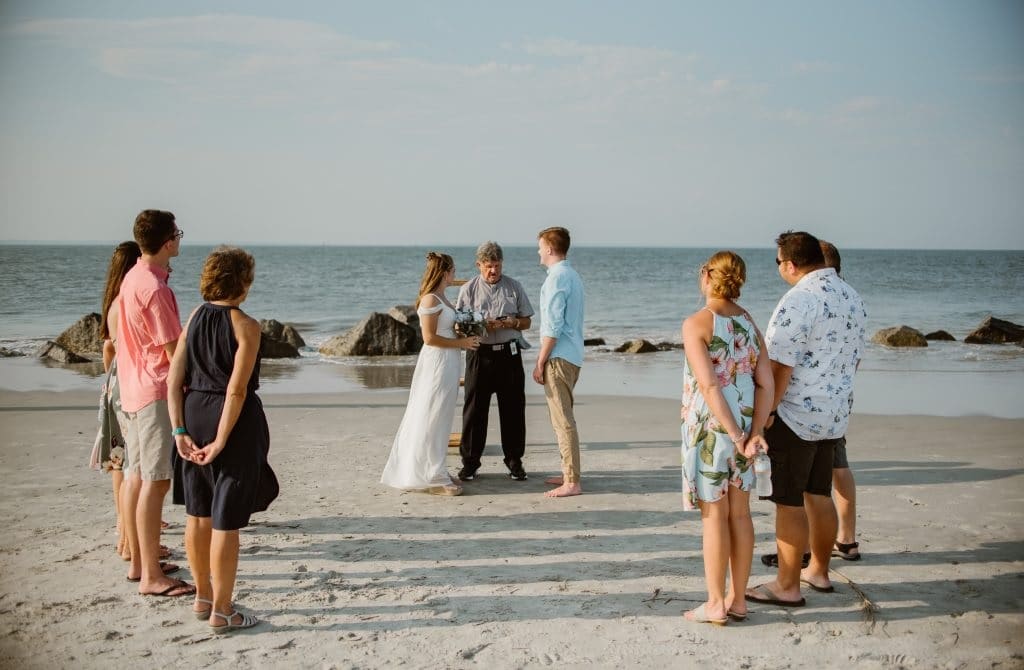 Couple stands barefoot in the sand on a beach with their officiant during their elopement ceremony, with their families' on either side of them.