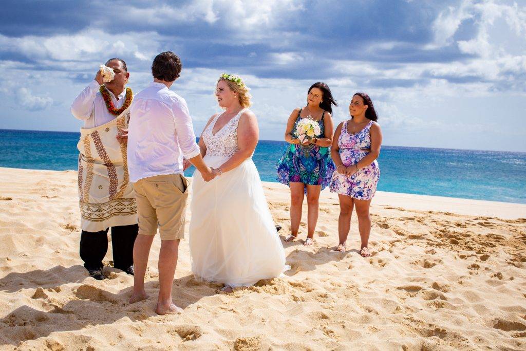 Couple stands hand in hand on a beach in Hawaii in front of their officiant as they blow into a ceremonial shell and two woman stand behind the bride smiling.