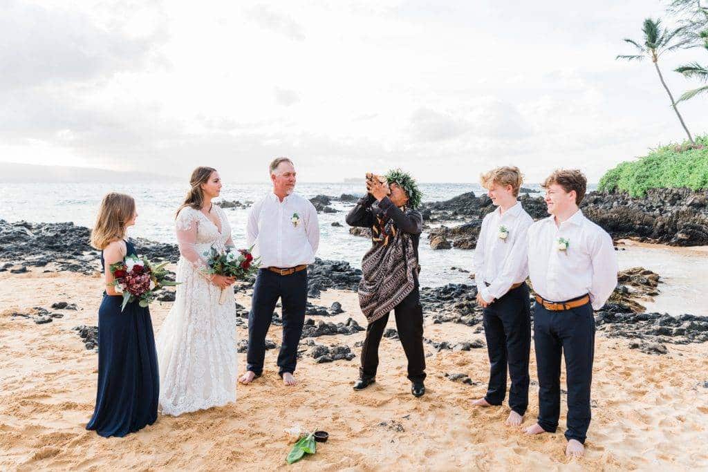 A couple stands beside their officiant, as he blows into a ceremonial shell during their Makena Cove elopement ceremony, with their kids on either side of them.