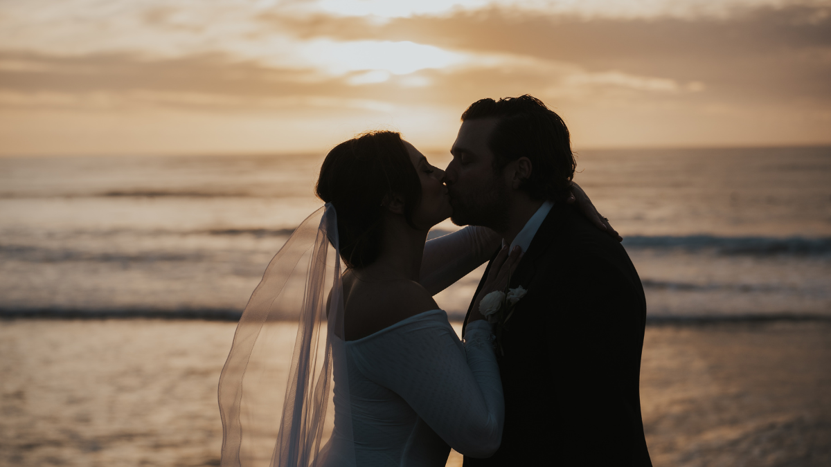 The dark silhouette of a bride and groom kissing on a sandy beach at sunset. The bride wears an off-the-shoulder wedding dress with a long, sheer veil catching the light, and the groom is dressed in a dark suit. The background shows gentle ocean waves rolling under a golden, cloudy sky with the sun setting over the water.