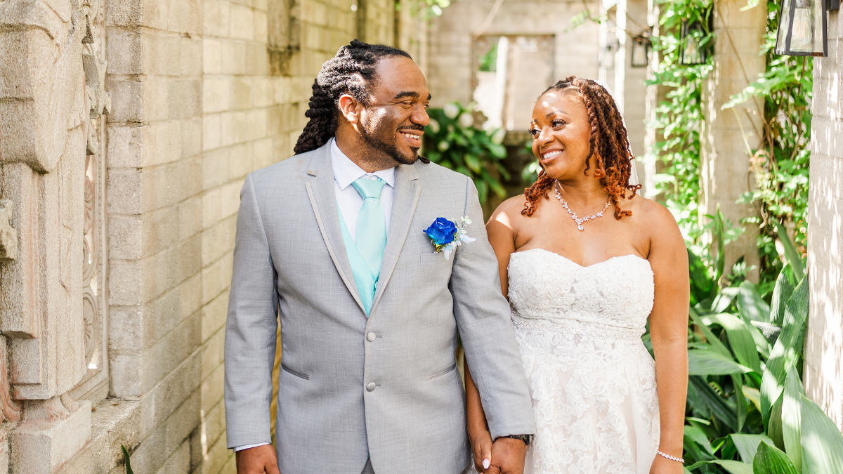 A Black bride and groom holding hands and smiling warmly at each other during an outdoor wedding ceremony. The groom features long locs pulled back, a light gray suit jacket, a mint blue tie and vest, and a vibrant royal blue rose boutonniere. The bride wears a strapless, sweetheart neckline lace wedding dress with her reddish-brown locs styled in a half-up look. They are standing in an intimate garden or courtyard alleyway lined with light-colored brick walls, stone carvings, and green tropical plants.