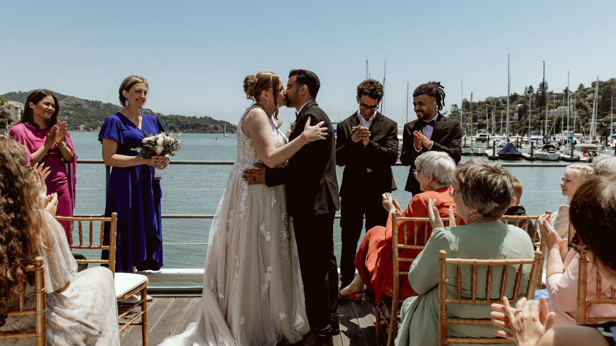 A modern elopement wedding ceremony takes place outdoors on a sunny wooden deck overlooking a marina with boats and green hills. A bride in an A-line lace wedding dress and a groom in a black suit share a first kiss as a small group of about ten wedding guests sitting in gold bamboo chairs and standing nearby clap and smile.
