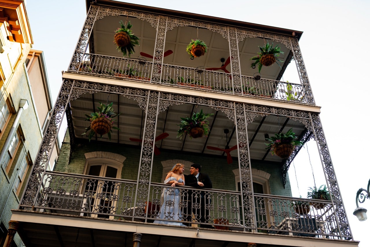 Couple dressed in formal wedding attire on a French Quarter balcony in New Orleans with hanging ferns and ornate iron railings.