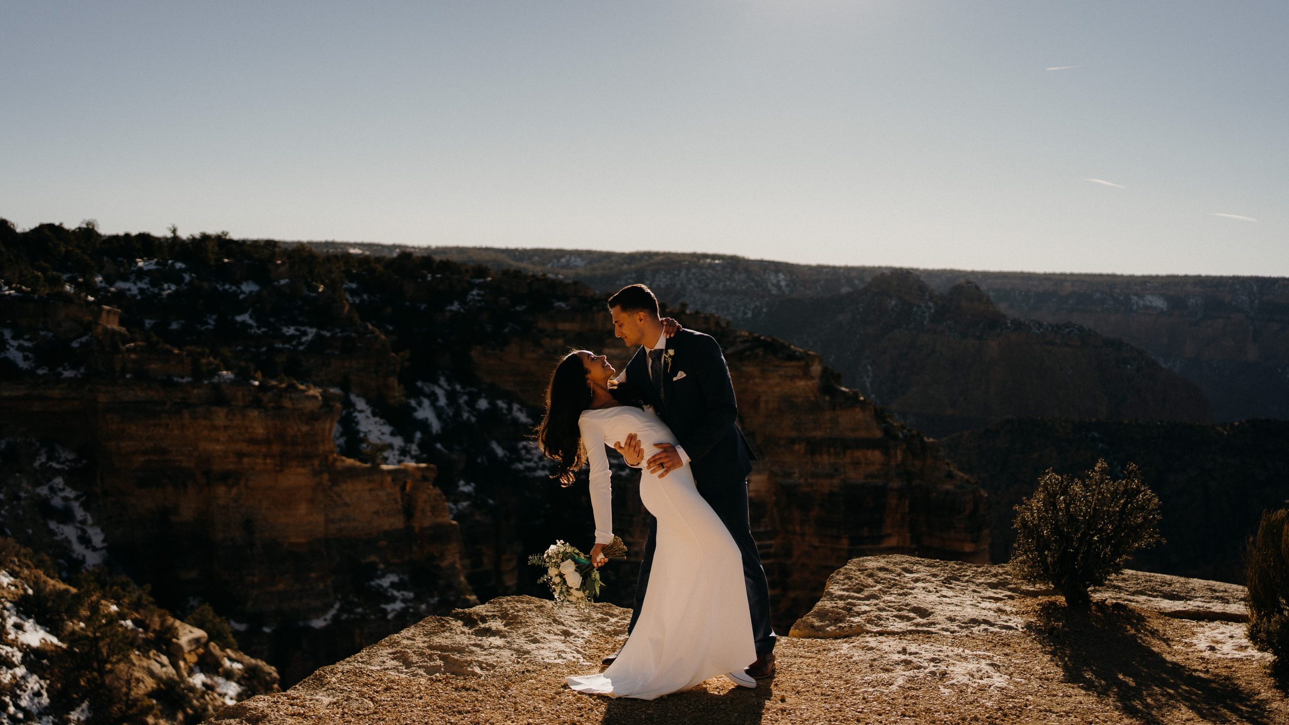 Bride and groom share a romantic dip and kiss during a grand canyon elopement at sunset, standing on a rocky cliff edge with layered canyon walls in the background.