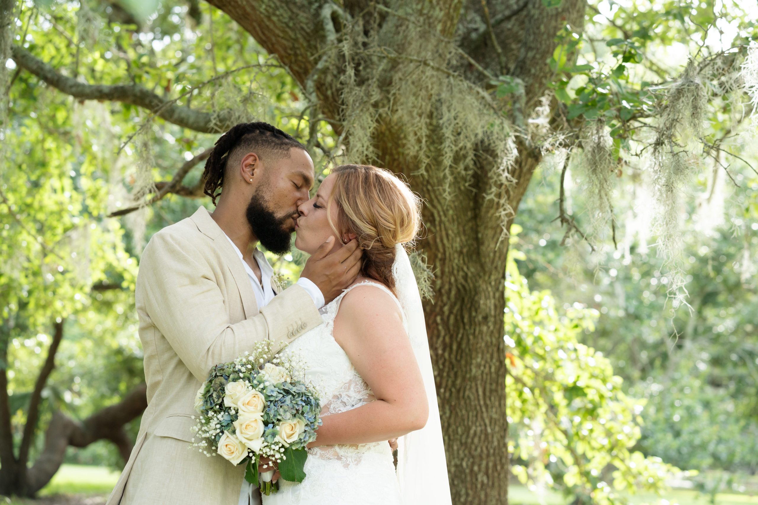 Bride and groom share a kiss during a garden elopement beneath a large oak tree draped with Spanish moss, holding a bouquet of white and blue flowers.
