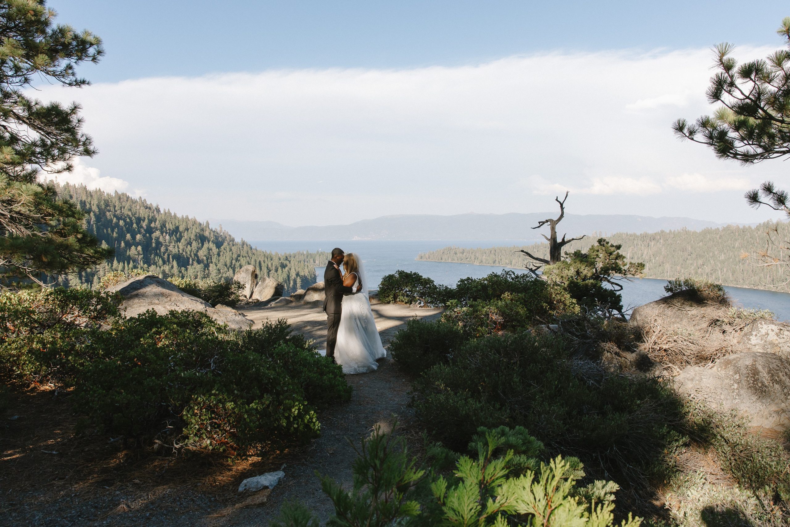 Bride and groom embrace during a scenic mountain elopement overlooking a blue lake, surrounded by pine trees, rocky terrain, and distant mountain ridges.