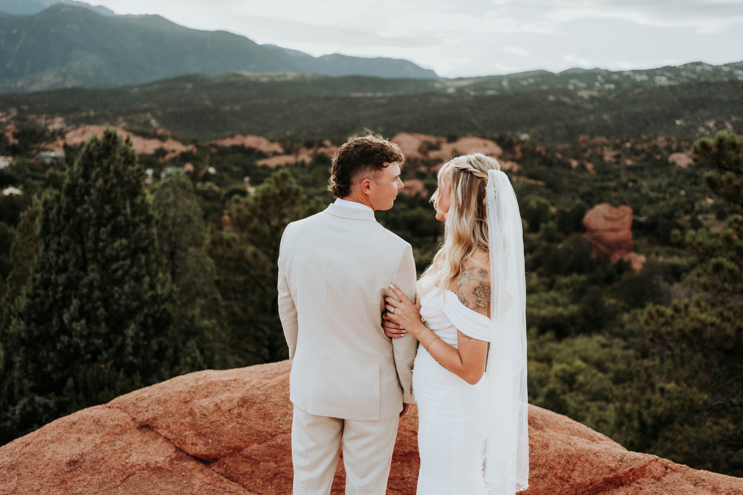 Bride and groom stand together on a red rock overlook during a Colorado elopement, looking out over a green valley with mountains in the distance.