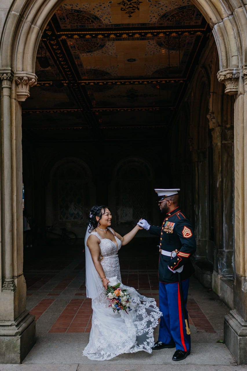 A bride in a lace dress and a groom in military uniform share a smile beneath stone arches, illustrating why do couples elope to find quiet, historic moments away from the crowd.