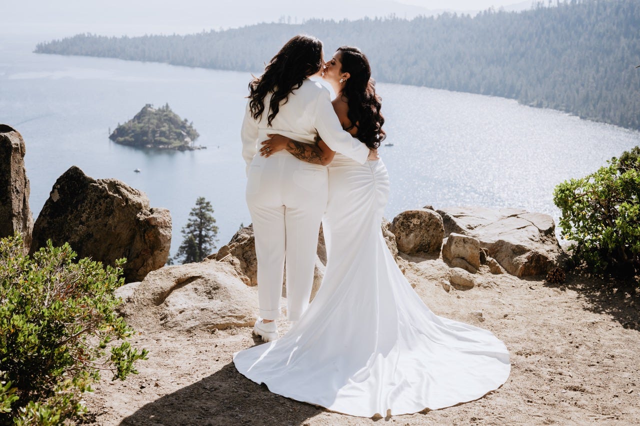 Two brides share a kiss during their mountain elopement, wearing white wedding outfits while standing on a rocky overlook above Emerald Bay in Lake Tahoe.