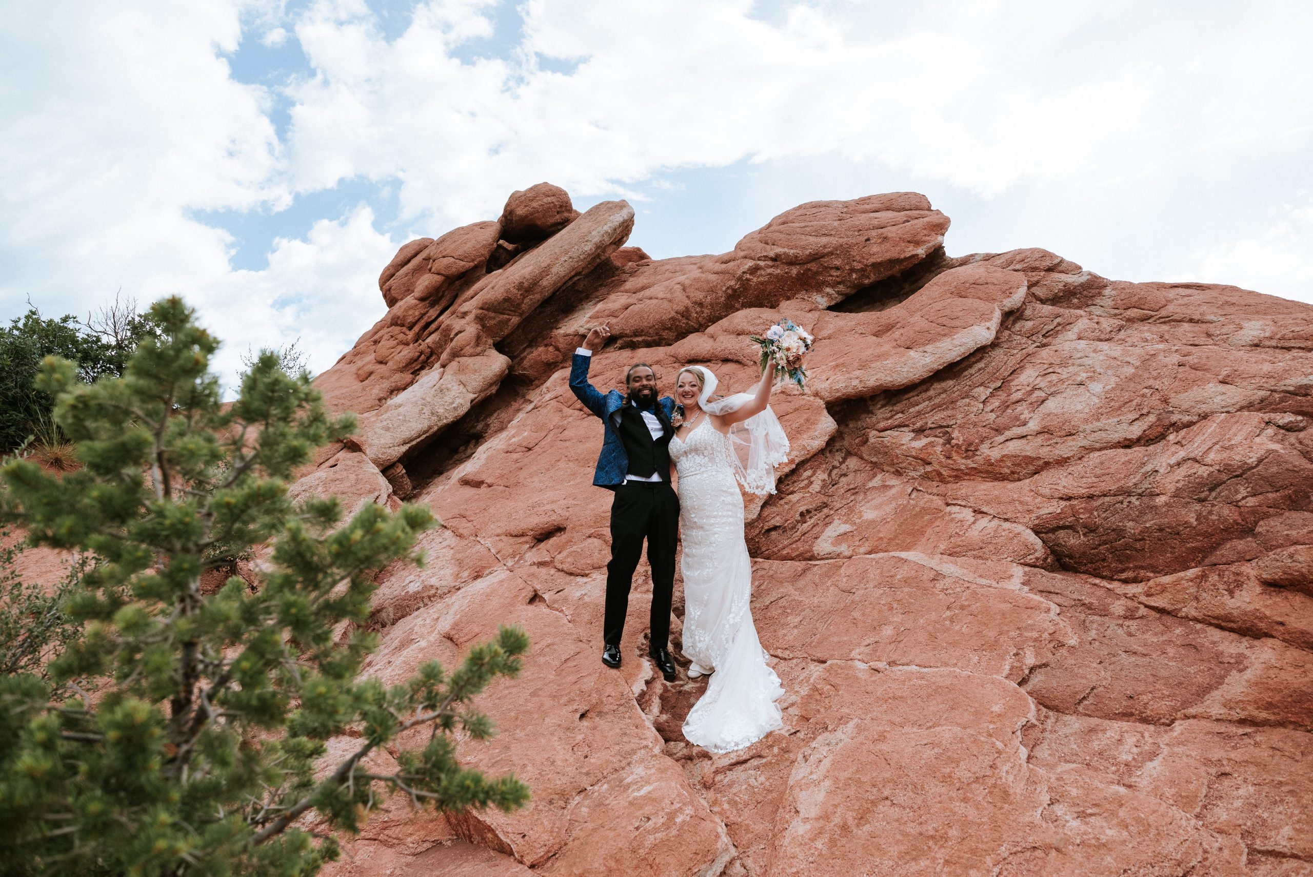 Newlyweds celebrate their Colorado elopement while standing on dramatic red rock formations, raising their arms and bouquet beneath a bright sky at a scenic outdoor location.