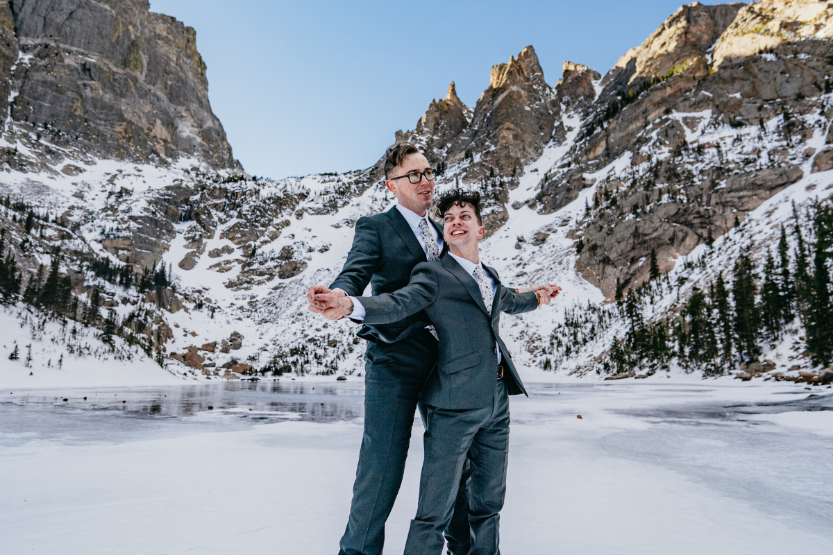 A wide shot of a couple in charcoal grey suits celebrating their winter wedding on a frozen, snow-covered lake. They are holding hands with arms outstretched in a joyful pose, surrounded by towering, snow-dusted rocky mountains under a clear blue sky.