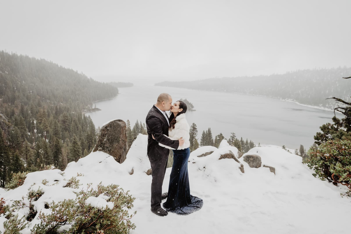 A couple shares a kiss on a snowy cliffside at the Emerald Bay overlook in Lake Tahoe. The woman wears a deep navy velvet gown and a white fur jacket, while the man is in a dark suit. Behind them, the misty grey lake and Fannette Island are framed by evergreen forests and snow-dusted rocks under an overcast sky.