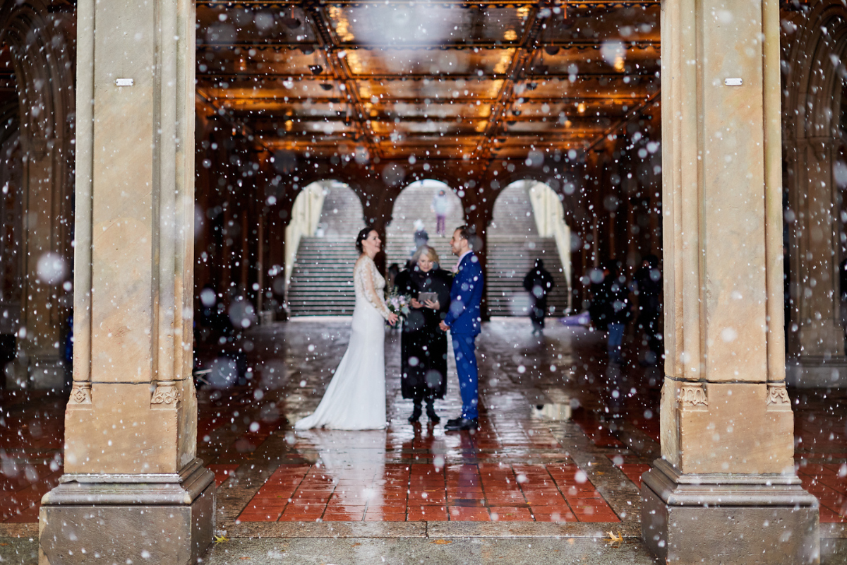 A bride in a long-sleeved lace gown and a groom in a bright blue suit stand before an officiant during a winter wedding ceremony inside the Bethesda Terrace Arcade in Central Park, NYC. Large, soft snowflakes or rain droplets are blurred in the foreground, creating a bokeh effect against the ornate tiled ceiling and sandstone arches.