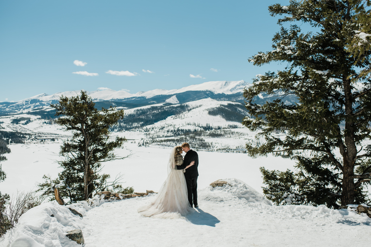 A romantic shot of a bride and groom embracing at the 3M Curve overlook. The bride wears a flowing white gown with a grey fur stole, and the groom is in a classic black suit. They stand on a snow-covered rocky ledge overlooking a massive, sun-drenched valley and distant white mountain peaks under a clear blue sky.