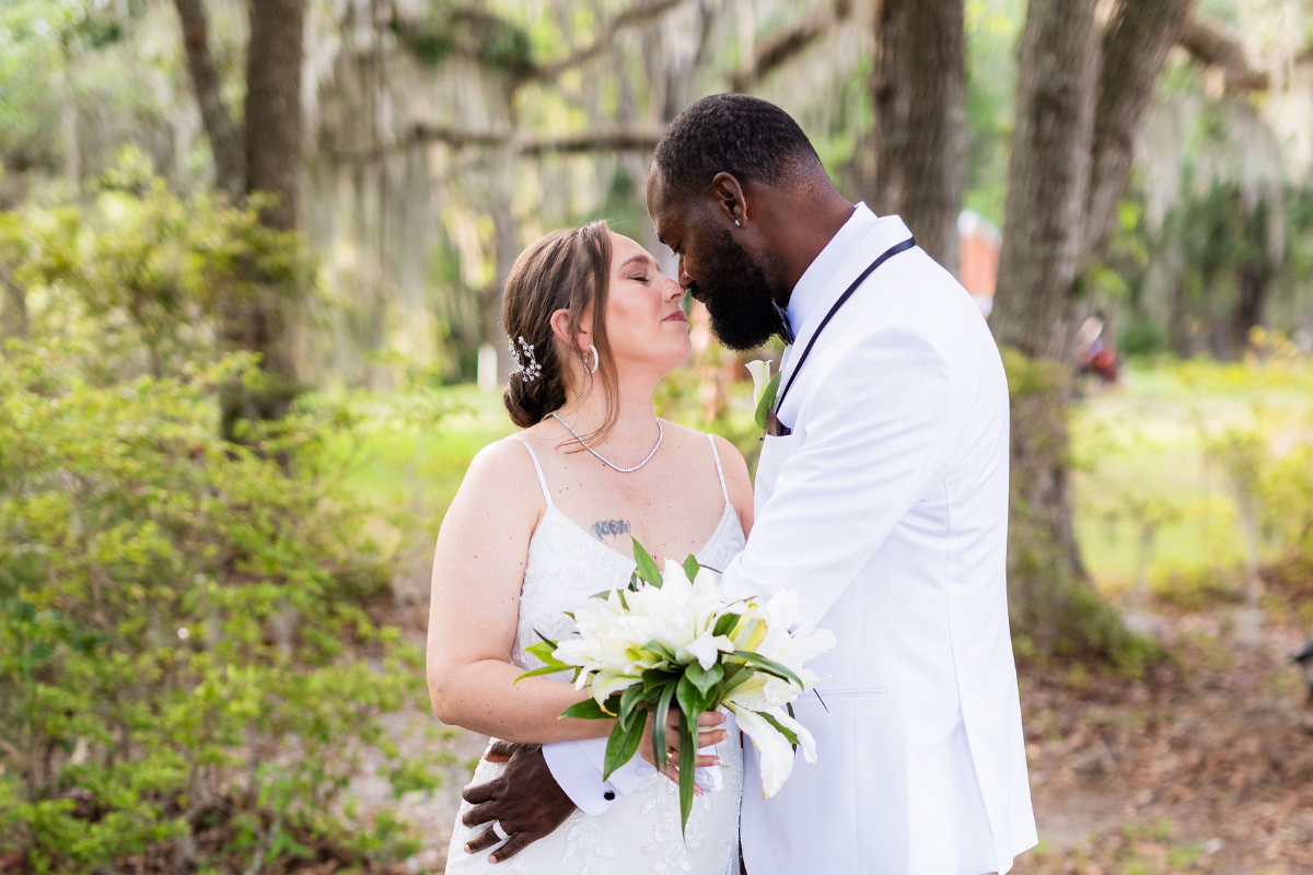 A medium shot of a wedding couple sharing an intimate moment outdoors in Charleston, South Carolina after their winter elopement. The bride, with her hair in an elegant updo, wears a white spaghetti-strap gown and holds a bouquet of white lilies. The groom, dressed in a sharp white tuxedo with black lapel piping, leans in close to touch his forehead against hers. They are surrounded by lush greenery and large trees with hanging Spanish moss in the soft-focus background.