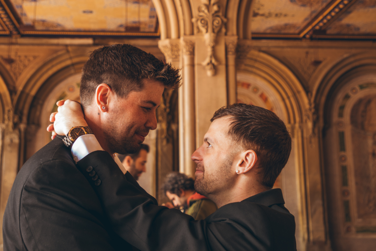 A close-up, profile shot of two men in dark suits embracing inside the Bethesda Terrace Arcade in Central Park after their winter elopement. One man has his arms wrapped around the other’s neck, showing a gold watch and a wedding ring. They are looking into each other's eyes with smiles. The background features the arcade’s iconic golden-toned Minton tile ceiling and ornate sandstone arches.