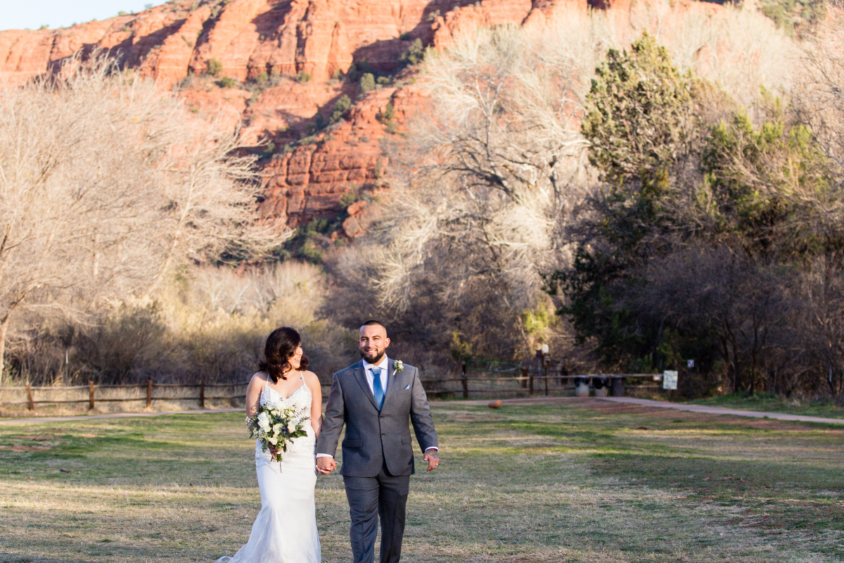 A wide shot of a wedding couple walking hand-in-hand through a grassy field at the Crescent Moon Day Use Area in Sedona, Arizona. The bride wears a fitted white lace gown and carries a bouquet of white flowers and greenery, looking toward the groom with a smile. The groom, in a grey suit with a bright blue tie, walks beside her looking toward the camera. In the background, towering orange-red rock formations rise behind a line of winter trees under a clear, late-afternoon sky.