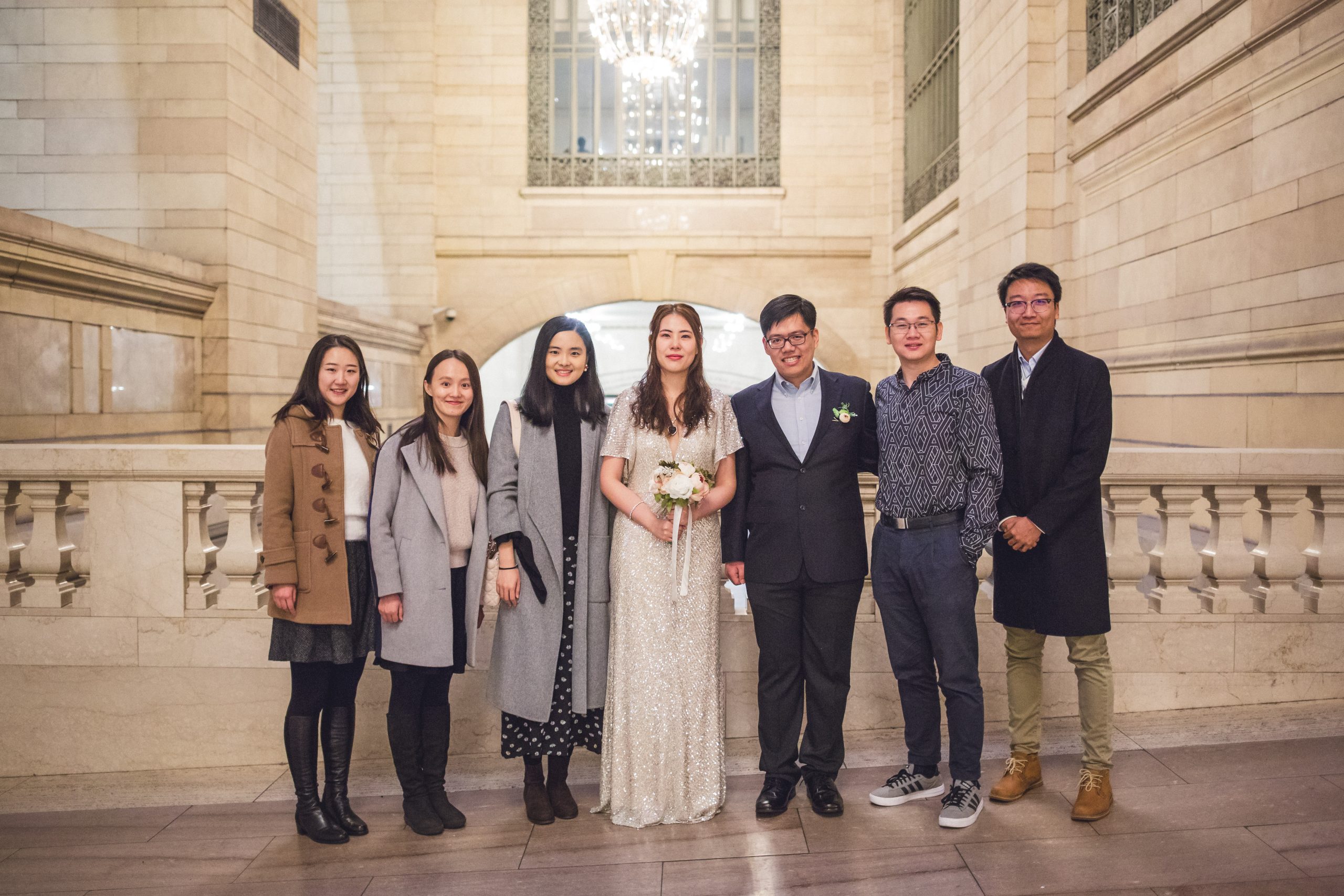 A group of wedding guests posing inside a grand, stone-walled New York City building, wearing stylish winter guest attire including wool overcoats, toggle coats, and boots.