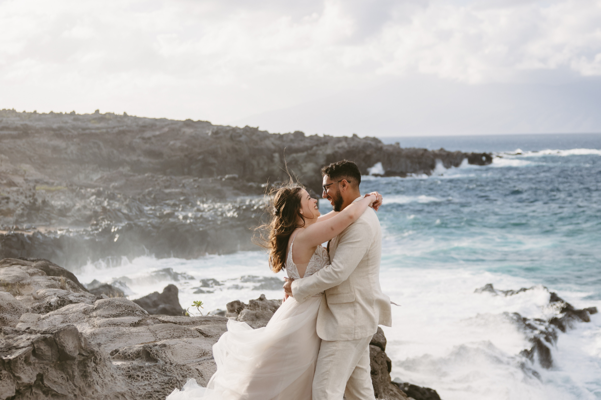 A joyous wedding couple embraces on a jagged, volcanic rock cliff at Ironwoods Beach in Maui. The bride, in a sleeveless white gown with a flowing skirt, has her arms wrapped around the groom’s neck, her hair blowing in the wind. The groom, wearing a light beige linen suit and glasses, holds her waist as they laugh together. In the background, white waves crash against the dark rocks under a bright, hazy sky.