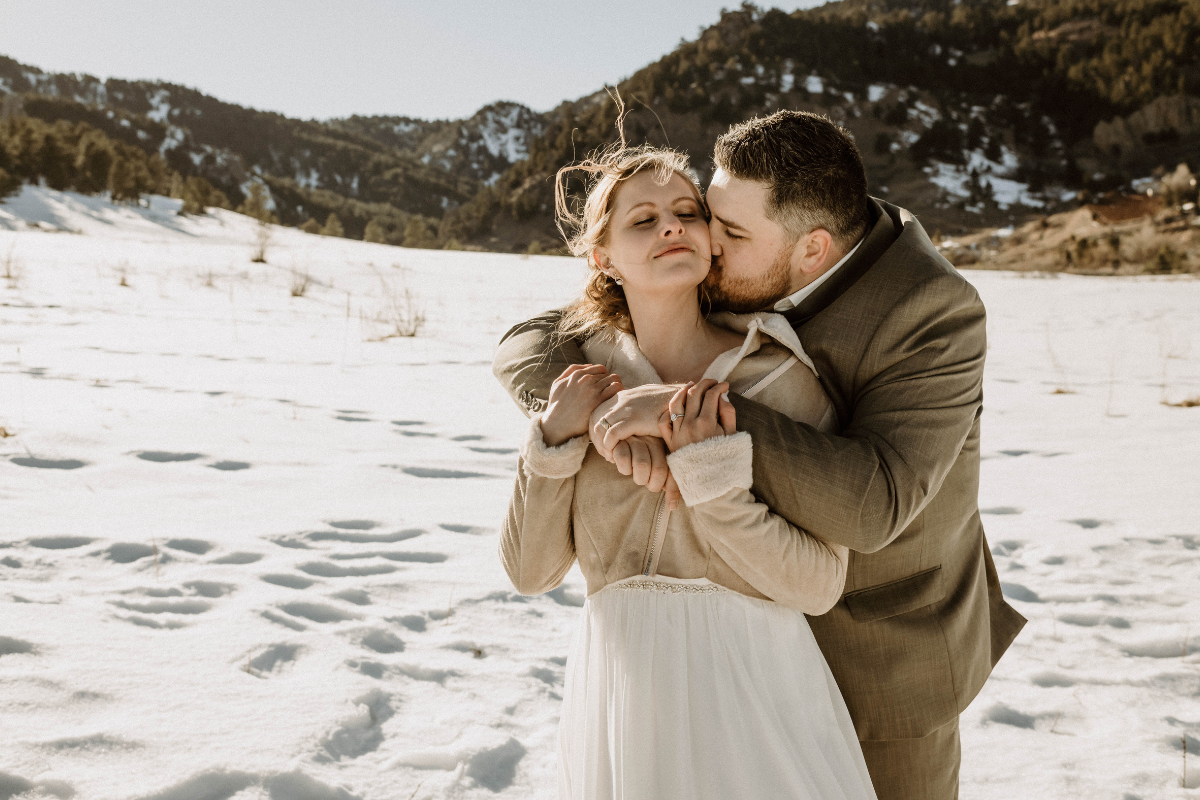 A close-up of a wedding couple embracing in a sunlit, snow-covered meadow at Moraine Park after their winter elopement. The groom, wearing a tan suit, stands behind the bride and kisses her cheek while wrapping his arms around her. The bride, wearing a white gown and a tan shearling-lined jacket, has her eyes closed with a peaceful expression. The background features a vast snowy field leading toward rolling pine-covered hills under a clear sky.