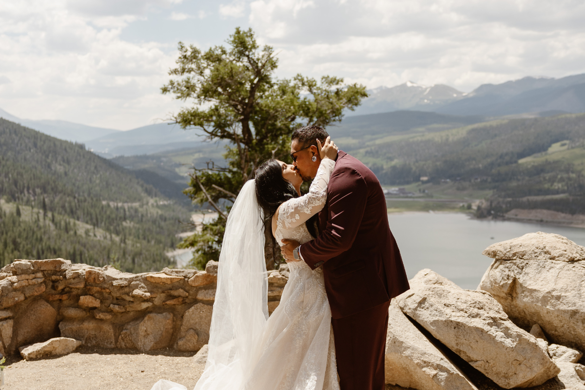 A wedding couple shares a romantic kiss at Sapphire Point Overlook. The bride, in a long-sleeved lace white gown with a flowing veil, leans back slightly as the groom, wearing a burgundy suit, holds her face. They are standing on a rocky ledge overlooking a vast valley that includes a large blue lake and rolling green mountains under a bright, partly cloudy sky.
