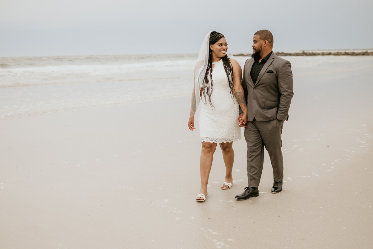 A wedding couple walks hand-in-hand along a wide, sandy beach in St. Johns County under a soft, overcast sky after their winter elopement. The bride wears a short white lace dress, a veil, and white sandals, while the groom wears a grey suit with a black shirt. They are looking at each other and smiling as they walk near the water's edge with gentle waves in the background.
