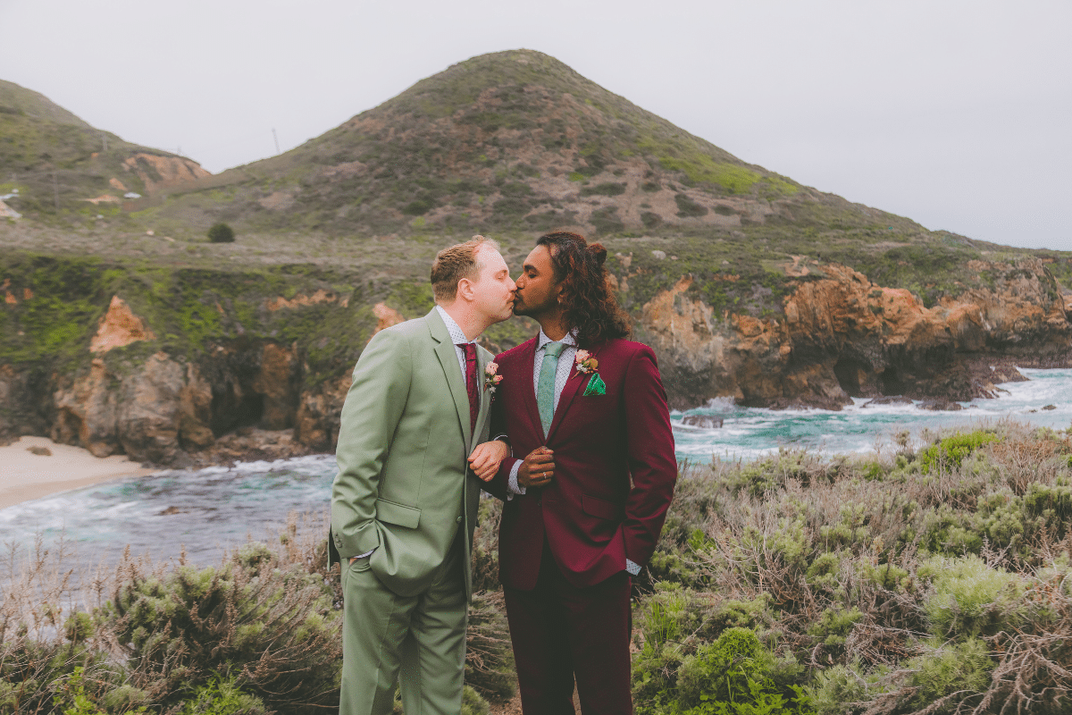A same-sex couple sharing a kiss during their big day on the Big Sur coast. One groom wears a sage green suit with a burgundy tie, and the other wears a burgundy suit with a sage green tie. They are standing on a cliff overlooking a rocky coastline and turquoise ocean under an overcast sky.