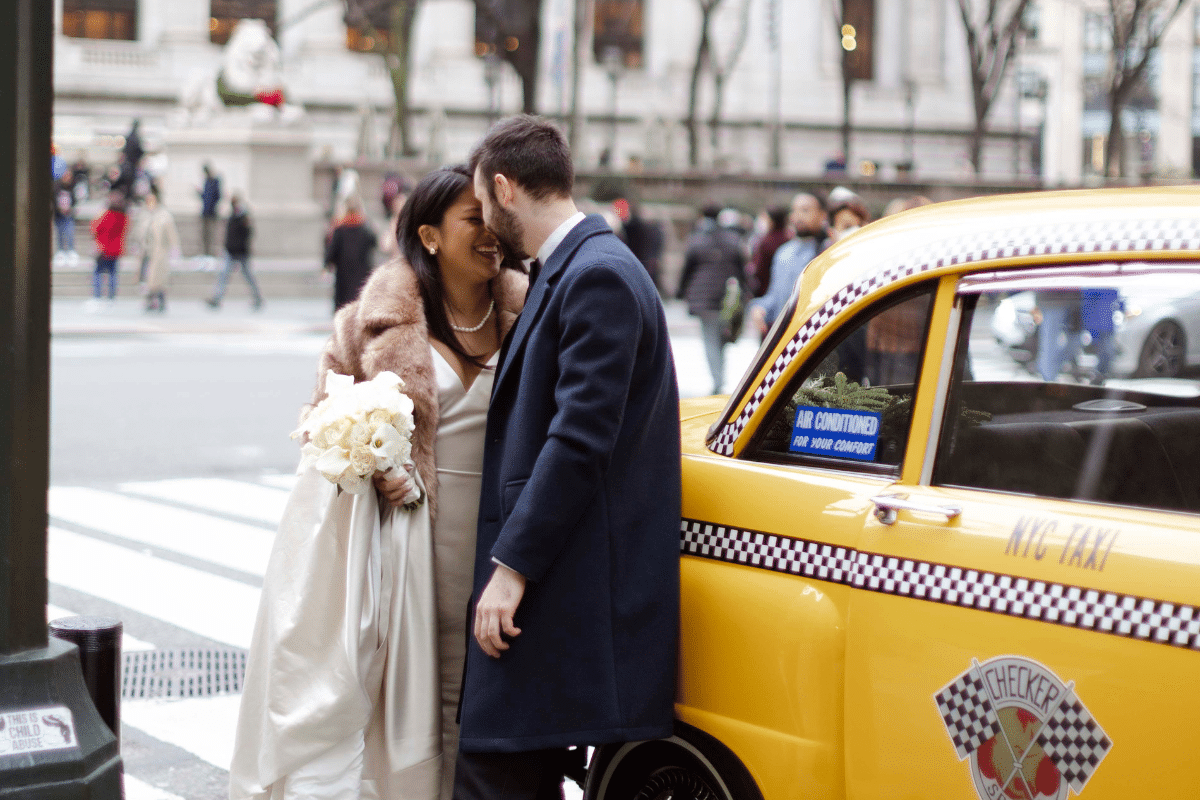 A bride and groom laughing together next to a vintage yellow Checker taxi in New York City during their urban elopement wedding. The bride wears a white gown with a fur stole and holds a white bouquet, while the groom wears a long navy overcoat.