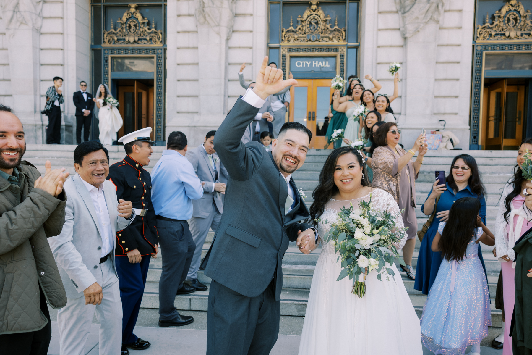A joyful bride and groom celebrating on the exterior steps of San Francisco City Hall. The groom, in a charcoal grey suit, makes a "shaka" hand gesture while the bride, in a long-sleeved lace gown, smiles while holding a white and green bouquet. They are surrounded by cheering family and friends, with the ornate gold-detailed doors of City Hall in the background.