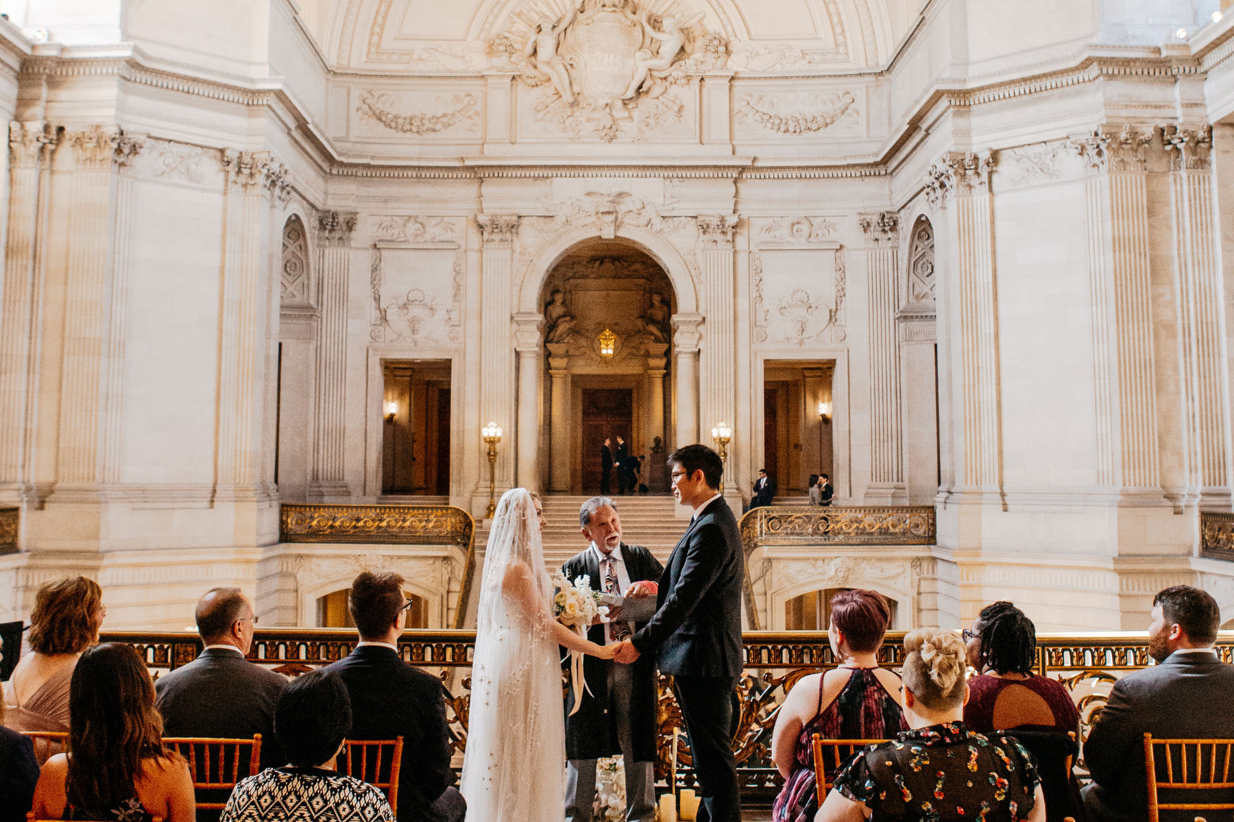 A medium shot from behind seated guests of a wedding ceremony on the fourth floor of the historic San Francisco City Hall. The bride in a beaded gown and the groom in a suit hold hands facing a male officiant on a balcony overlooking the rotunda. The elaborate limestone architecture, gold railings, and ornate reliefs of the Beaux-Arts building are visible in the background.
